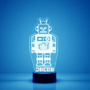 以下が含まれることがあります： 黒いベースが付いた白いロボットのナイトライト。ロボットは、目、口、鼻のついた顔をしています。ロボットには「JACOB」と書かれたネームプレートが付いています。