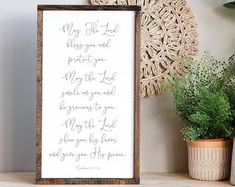 May include: A white wooden sign with a brown frame features a black calligraphy quote about blessings and peace. The quote reads "May The Lord bless you and protect you. May the Lord smile on you and be gracious to you. May the Lord show you his favor and give you His peace." The quote is attributed to Numbers 6:24-26.
