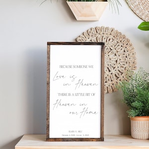May include: A wooden sign with a white background and black text. The text reads "Because someone we love is in Heaven, there is a little bit of Heaven in our home." The sign also includes the name "Jamie F. Bell" and the dates "December 8, 1989 - October 5, 2021".