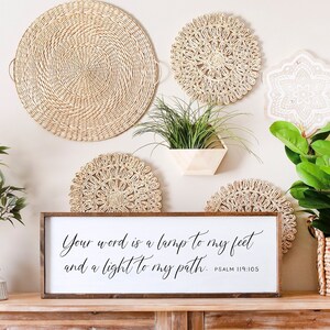 May include: A wooden sign with the text "Your word is a lamp to my feet and a light to my path. Psalm 119:105" in black calligraphy. The sign is hanging on a wall with woven wall decor and potted plants.