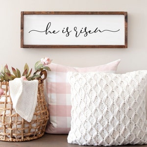 Puede incluir: Un letrero de madera con el texto "He is risen" en escritura cursiva negra. El letrero est&aacute; colgado en una pared blanca sobre una almohada a cuadros rosa y blanca y una almohada de punto blanco.