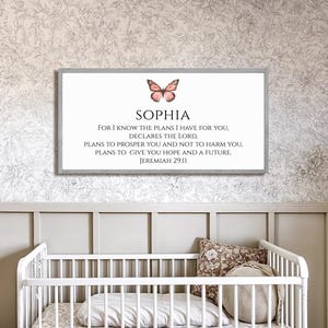 May include: A framed print featuring the name "SOPHIA" and a butterfly illustration, with a Bible verse from Jeremiah 29:11. The artwork is displayed above a white cot in a nursery setting, with a floral wallpaper background.
