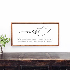 May include: A rectangular wooden framed sign with the word "nest" in cursive script. Below the word is a definition: "(N.) A snug, comfortable or cozy residence; a retreat, refuge or resting place; home."