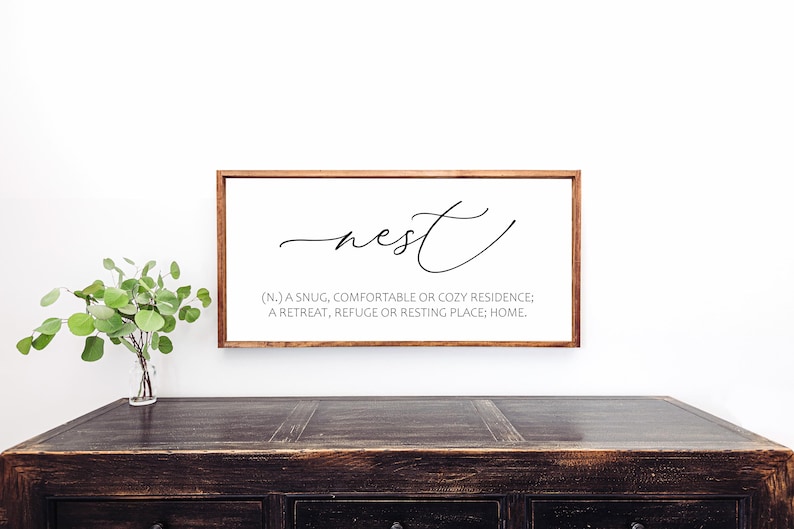 May include: A rectangular wooden framed sign with the word "nest" in cursive script. Below the word is a definition: "(N.) A snug, comfortable or cozy residence; a retreat, refuge or resting place; home."
