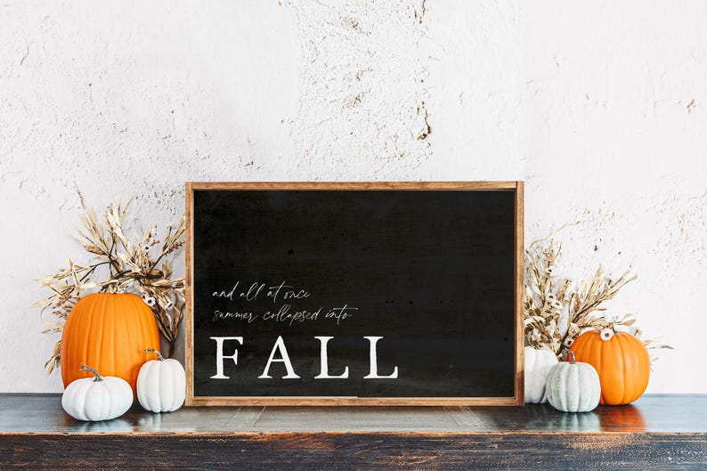 May include: A black framed sign with the word "FALL" in white, along with the phrase "and all at once summer collapsed into". The sign is surrounded by pumpkins and dried foliage, creating a fall theme.