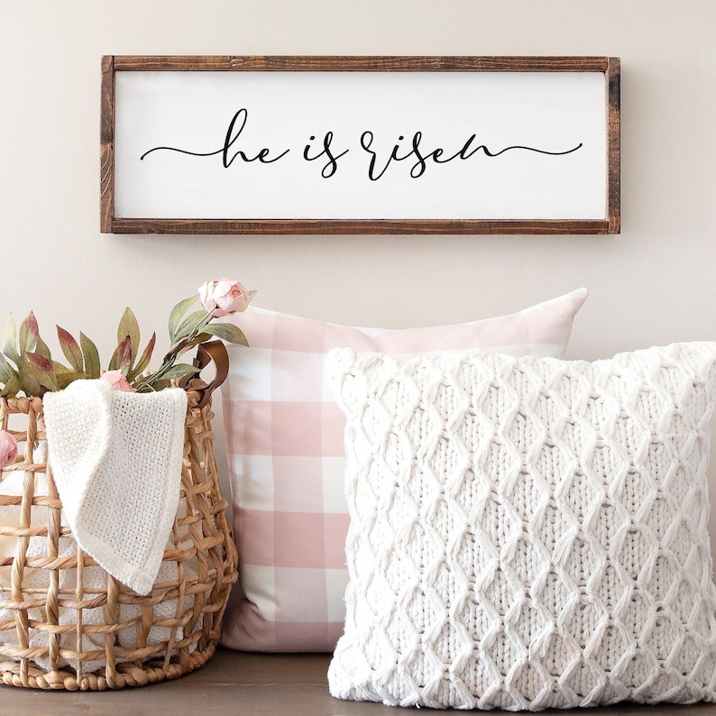 Puede incluir: Un letrero de madera con el texto "He is risen" en escritura cursiva negra. El letrero est&aacute; colgado en una pared blanca sobre una almohada a cuadros rosa y blanca y una almohada de punto blanco.