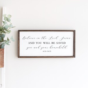 May include: A wooden sign with a white background and brown frame. The sign features a black cursive quote: "Believe in the Lord Jesus. AND YOU WILL BE SAVED you and your household." The quote is followed by "ACTS 16:31".