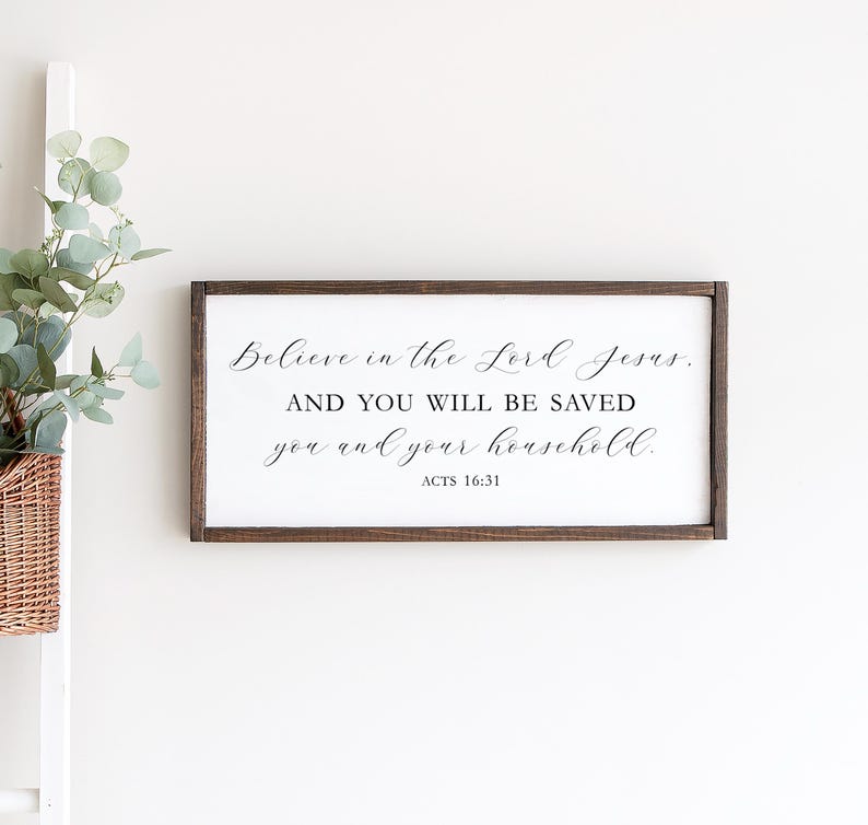 May include: A wooden sign with a white background and brown frame. The sign features a black cursive quote: "Believe in the Lord Jesus. AND YOU WILL BE SAVED you and your household." The quote is followed by "ACTS 16:31".
