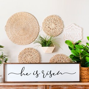 Puede incluir: Un letrero de madera con el texto "He is risen" en escritura cursiva negra. El letrero se muestra en un estante de madera con una variedad de decoraci&oacute;n de pared tejida y plantas en macetas.