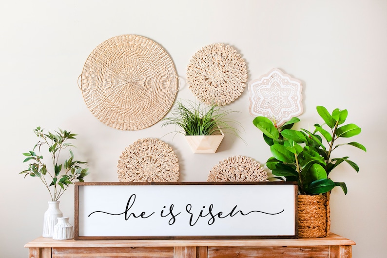 Puede incluir: Un letrero de madera con el texto "He is risen" en escritura cursiva negra. El letrero se muestra en un estante de madera con una variedad de decoraci&oacute;n de pared tejida y plantas en macetas.
