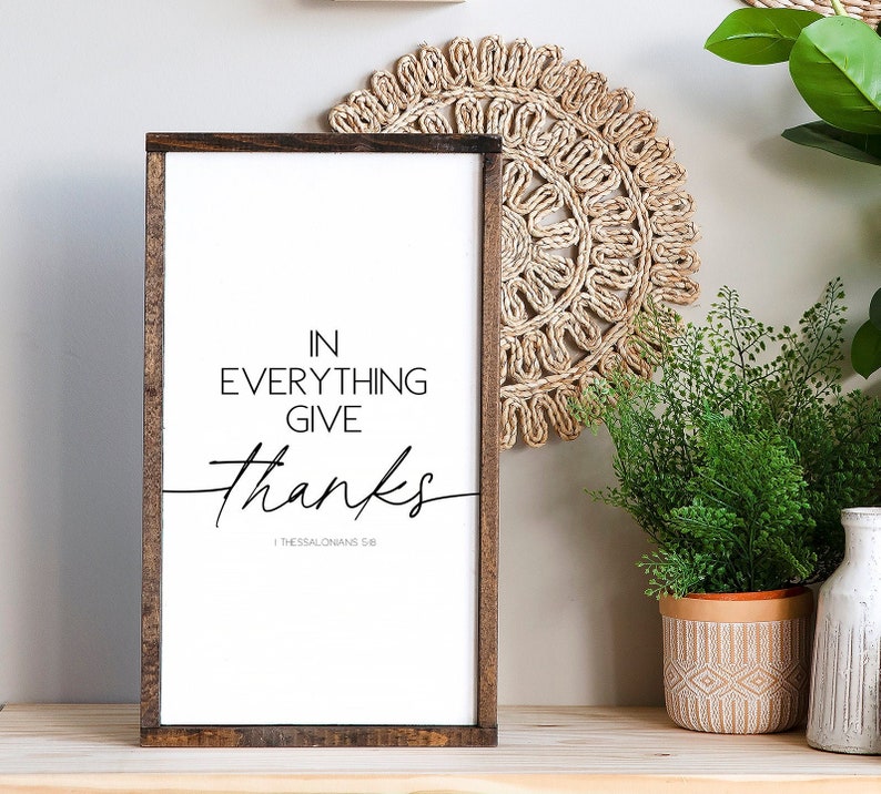 Op de afbeelding: Een bruin houten geframed bord met een witte achtergrond en zwarte tekst die "In alles dankbaarheid 1 Thessalonicenzen 5:18" luidt.