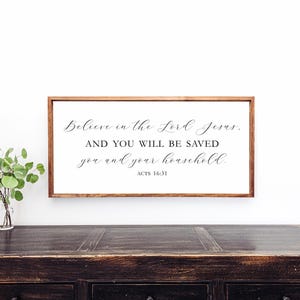 May include: A wooden sign with a white background and a brown frame. The sign has a black cursive quote that reads "Believe in the Lord Jesus. AND YOU WILL BE SAVED you and your household. ACTS 16:31"