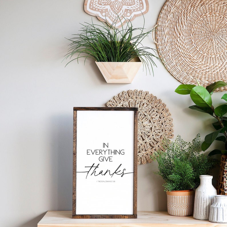 Op de afbeelding: Een ingelijst houten bord met een witte achtergrond en zwarte tekst die "In alles dankbaarheid 1 Thessalonicenzen 5:18" luidt. Het bord staat op een houten tafel met een potplant en een geweven mat.