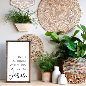 May include: A wooden sign with the text "In the morning when I rise give me Jesus." The sign is hanging on a wall with a variety of plants and woven wall decor.