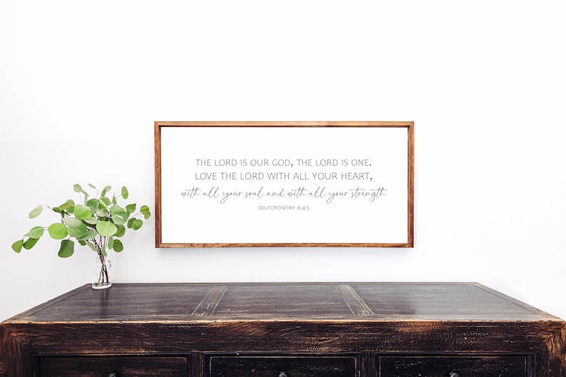 May include: A framed print with a white background and black text. The text reads "THE LORD IS OUR GOD, THE LORD IS ONE. LOVE THE LORD WITH ALL YOUR HEART, with all your soul and with all your strength DEUTORONOMY 6:4-5". The frame is brown wood.