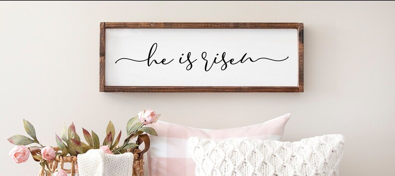 Puede incluir: Un letrero de madera con el texto "He is risen" en escritura cursiva negra. El letrero est&aacute; colgado en una pared blanca con una almohada a cuadros rosa y blanca y una almohada de punto blanco delante.
