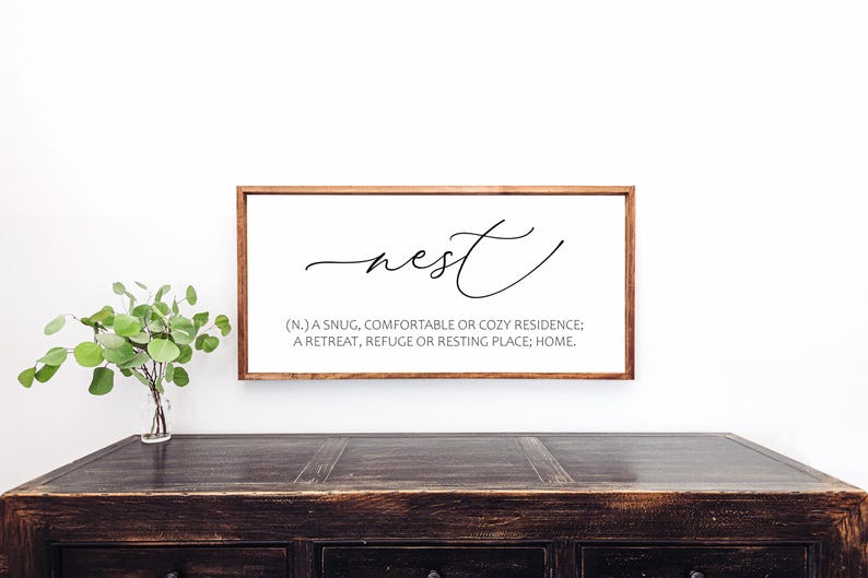 May include: A rectangular wooden framed sign with the word "nest" in cursive script. Below the word is a definition: "(N.) A snug, comfortable or cozy residence; a retreat, refuge or resting place; home."