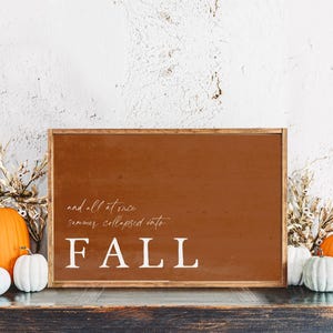 May include: A framed sign with the word "FALL" in white, with the phrase "and all at once summer collapsed into" above it. The sign is surrounded by pumpkins and dried foliage, creating a fall-themed display.