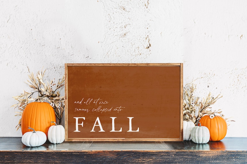 May include: A framed sign with the word "FALL" in white, with the phrase "and all at once summer collapsed into" above it. The sign is surrounded by pumpkins and dried foliage, creating a fall-themed display.