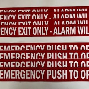 May include: Red rectangular signs with white text. The top signs read "EMERGENCY EXIT ONLY - ALARM WILL SOUND." The bottom signs read "IN EMERGENCY PUSH TO OPEN."