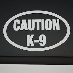 Könnte beinhalten: Weißer ovaler Aufkleber mit dem Text "CAUTION K-9" auf schwarzem Hintergrund.