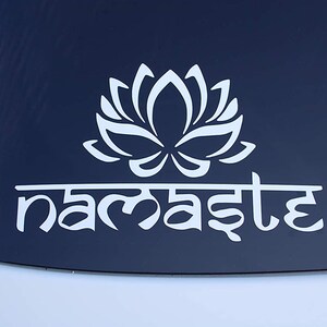 Puede incluir: Pegatina de vinilo blanco con una flor de loto estilizada y la palabra "namaste" en escritura sánscrita.
