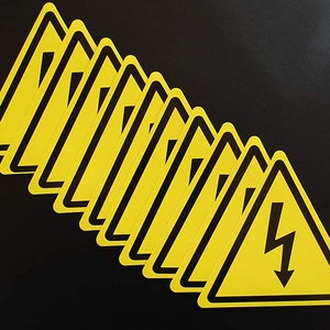 May include: A stack of ten yellow warning signs with black borders and a lightning bolt symbol. The signs are arranged in a fan shape.