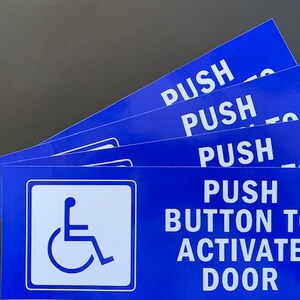 May include: Stack of blue signs with white text that reads "Push Button to Activate Door". The top sign has a wheelchair accessibility symbol.