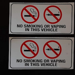 May include: Two white rectangular signs with black borders. Each sign has a red circle with a diagonal line through it. The top circle shows a cigarette with smoke coming from it. The bottom circle shows an electronic cigarette. The text on the signs reads "NO SMOKING OR VAPING IN THIS VEHICLE".