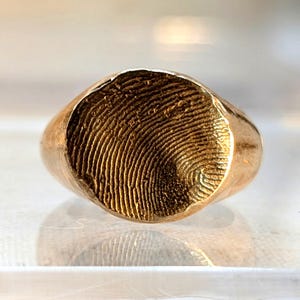 May include: A gold signet ring with a fingerprint design. The ring is a classic style with a rounded top and a smooth band.