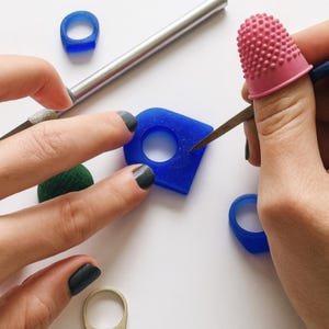 Puede incluir: Un dedal rosa texturizado, una herramienta de metal y varios anillos de resina azul están dispuestos sobre una superficie. Un anillo de resina azul tiene una forma geométrica con un orificio central, mientras que otros son circulares.
