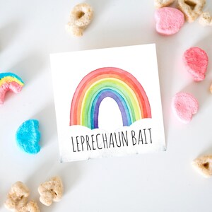 May include: A square card with a watercolor rainbow design and the words "LEPRECHAUN BAIT" in black text. Colorful marshmallow cereal pieces and other cereal pieces are scattered around the card on a white surface.