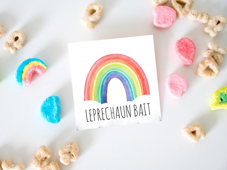 May include: A square card with a watercolor rainbow design and the words "LEPRECHAUN BAIT" in black text. Colorful marshmallow cereal pieces and other cereal pieces are scattered around the card on a white surface.