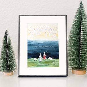 May include: Framed watercolor print with a religious theme. The artwork depicts angels, shepherds, and sheep, with the text "Glory to God in the Highest, and on earth peace, good will toward men." Two small green Christmas trees flank the frame.