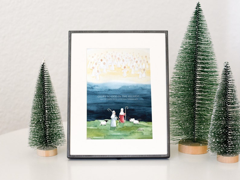 May include: Framed watercolor print with a religious theme. The artwork depicts angels, shepherds, and sheep, with the text "Glory to God in the Highest, and on earth peace, good will toward men." Two small green Christmas trees flank the frame.