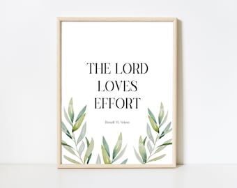 May include: A framed wall art print featuring the black text THE LORD LOVES EFFORT and Russell M. Nelson in a serif font on a white background, decorated with watercolour-style green and olive leaves along the bottom edges in a light wood frame.
