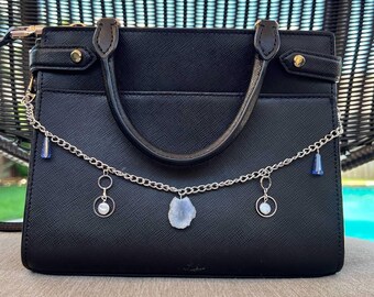 May include: Black handbag with a top handle and a decorative chain. The chain features blue and white charms, including a teardrop-shaped stone. The bag has gold-colored hardware and a structured design.