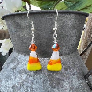 May include: A pair of dangle earrings featuring a candy corn design with white, orange, and yellow glass. The earrings have silver hooks and crystal accents.
