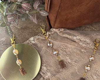 May include: Two gold-colored keychains with decorative charms. Each keychain features a series of clear and gold-colored beads, and a small brown gummy bear charm. The keychains are displayed on a gold-colored surface and a tan lace fabric.