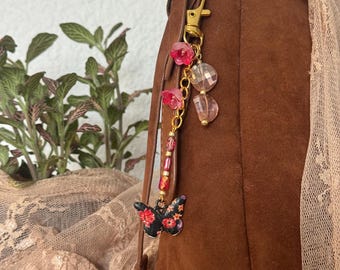 May include: A brown suede handbag with a gold clasp and a decorative charm. The charm features a black butterfly with a floral design, pink beads, and a gold chain. The background includes a plant and lace fabric.