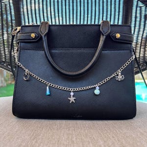 May include: Black handbag with a top handle and a silver chain. The chain is adorned with charms, including a starfish, a seahorse, and blue and white beads. The bag has gold-colored hardware.