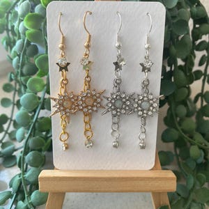 Könnte beinhalten: Zwei Paar Ohrhänger auf einer weißen Karte. Ein Paar ist goldfarben mit Stern- und Sonnenstrahl-Designs, das andere ist silberfarben mit ähnlichen Designs. Beide Paare haben kleine, baumelnde Akzente und werden auf einer kleinen Holzstaffelei präsentiert.