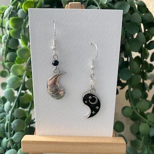 Könnte beinhalten: Ein Paar nicht übereinstimmender Ohrringe. Ein Ohrring zeigt einen silbernen Halbmond mit einem Sonnendesign, der andere einen schwarzen Halbmond mit Sternen und einem Mond. Beide Ohrringe haben silberne Haken und Perlenakzente.