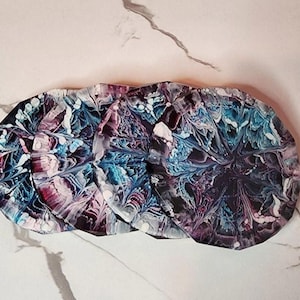 May include: Set of four round coasters with an abstract design. The coasters feature a swirling pattern of blue, purple, and white paint. The coasters are made of a glossy material and are arranged in a row.