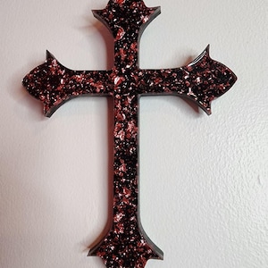 May include: A decorative cross with a dark frame and a textured, reddish-brown interior. The cross has a traditional shape with flared ends and a pointed base, suitable for wall display.
