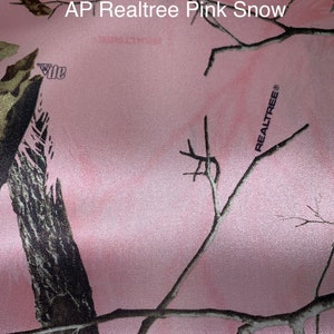 May include: Pink fabric with a camouflage pattern featuring branches and leaves. The fabric has the text "AP Realtree Pink Snow" and the Realtree logo.