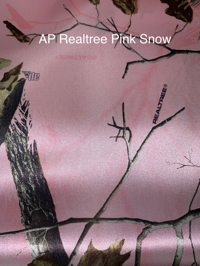 May include: Pink fabric with a camouflage pattern featuring branches and leaves. The fabric has the text "AP Realtree Pink Snow" and the Realtree logo.