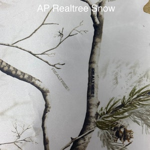May include: AP RealTree Snow camouflage pattern featuring a winter forest scene with bare tree branches, pine needles, and a pine cone. The design is predominantly white and gray, with subtle brown and green accents. The RealTree logo is visible.