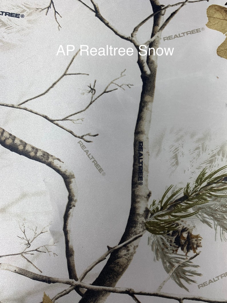 May include: AP RealTree Snow camouflage pattern featuring a winter forest scene with bare tree branches, pine needles, and a pine cone. The design is predominantly white and gray, with subtle brown and green accents. The RealTree logo is visible.