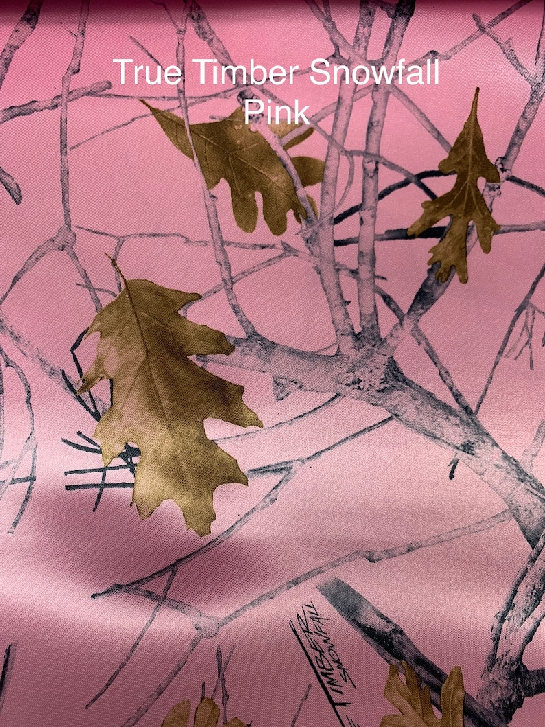 May include: Pink camouflage fabric with a pattern of tree branches and leaves. The fabric has the text "True Timber Snowfall Pink" printed on it. The leaves are a golden brown color.